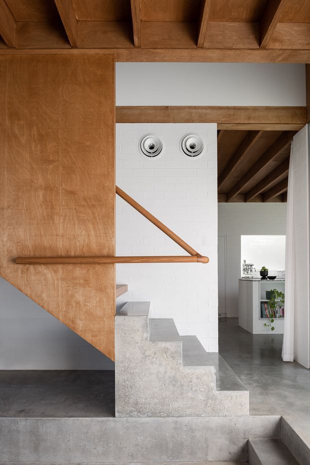 Interior architectural photograph focused on a sculptural staircase. A warm timber-clad stair and balustrade descend beside a white painted brick wall with circular wall lights, while exposed timber ceiling beams, polished concrete floors, and raw concrete stair treads create a restrained material palette. A partial open-plan room with built-in shelving is visible beyond.