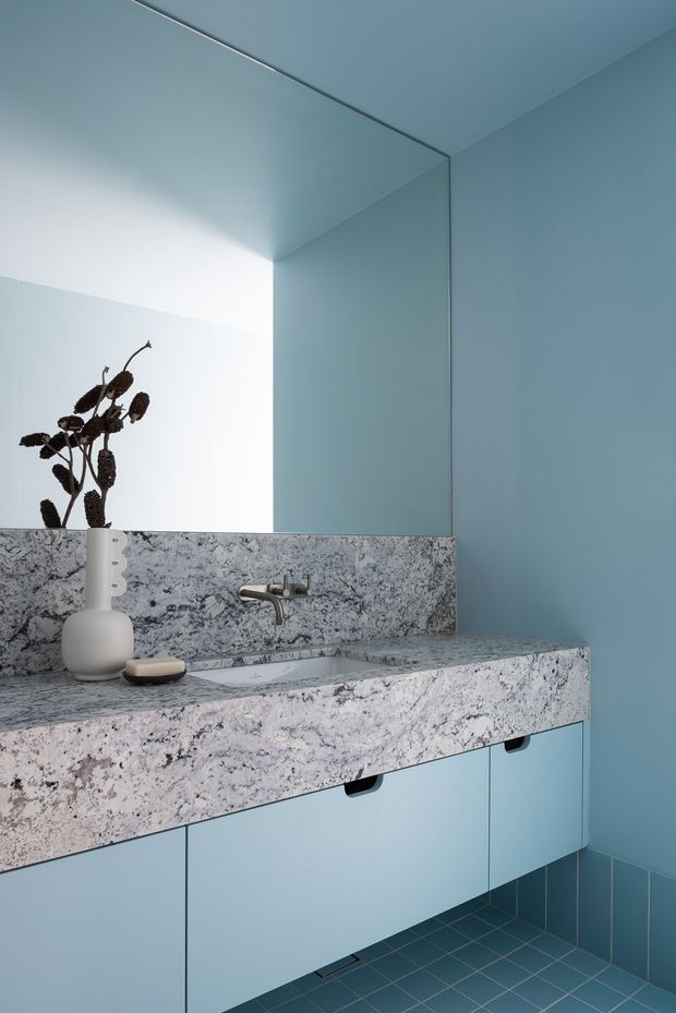 A compact, highly coordinated bathroom vanity in cool blue tones. The space features a thick speckled natural-stone benchtop and integrated basin, matching stone splashback, pale blue wall cabinetry with minimal black pulls, blue tiled flooring, and a large frameless mirror. A sculptural white vessel and dark dried botanical arrangement add restrained decoration.