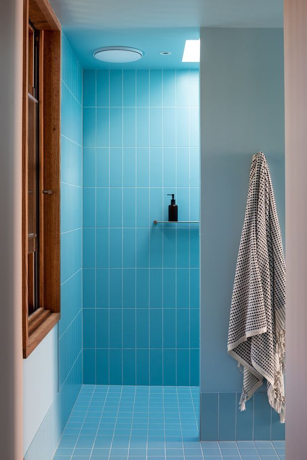 A compact modern shower room with vivid turquoise-blue ceramic tile covering the shower walls and floor. The space includes a frameless glass shower screen, a slim wall-mounted shelf with dark soap dispenser, a wood-framed window, recessed and circular ceiling lighting, and a patterned towel hanging on the right wall. The design is clean, graphic, and colour-focused with strong blue monochrome styling.