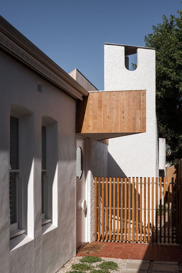 A narrow residential exterior passage or courtyard with white textured stucco walls, deep-set windows, warm timber cladding, and a projecting timber-clad upper volume. A tall white rendered architectural feature with a roof opening rises above the courtyard, while a vertical timber slat gate encloses the brick-paved area. Strong sunlight creates crisp shadows, with mature greenery at the edge of the frame.