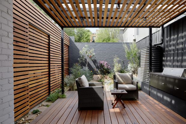 A compact, private outdoor courtyard terrace with a timber deck, slatted timber privacy fencing, and a timber pergola overhead. Dark painted masonry walls and black built-in cabinetry with a stainless-steel barbecue line one side, while outdoor lounge chairs and planting create a sheltered entertaining area.