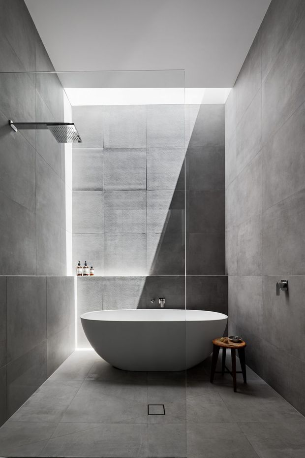 A minimalist contemporary bathroom with large-format grey concrete-look tiles covering the walls and floor. A freestanding white oval bathtub sits behind a clear frameless glass shower screen, with a ceiling-mounted rain shower on the left. A narrow skylight or clerestory opening creates a strong diagonal beam of natural light across the tiled back wall. The space is sparse and monochromatic, with a small wooden stool beside the bath.