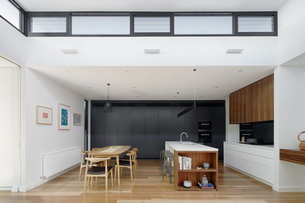 Bright contemporary open-plan kitchen and dining area with a double-height ceiling, high clerestory windows, pale timber flooring, white walls, and dark matte cabinetry. A central island with a light benchtop and timber shelving separates the kitchen from a timber dining table and chairs. Vertical timber joinery adds warmth to the kitchen wall, while framed artwork and minimalist lighting provide restrained decoration.