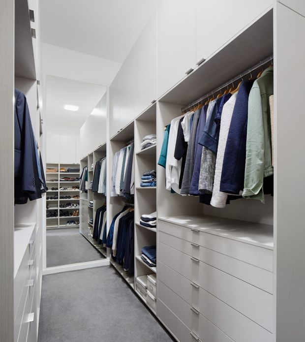 A bright, narrow walk-in wardrobe with floor-to-ceiling white built-in cabinetry, open hanging rails, shelves, and multiple drawers. Clothing is neatly organised in predominantly blue, grey, white, and muted green tones. A full-height mirror at the end of the corridor reflects additional storage, while grey carpet covers the floor and recessed ceiling lighting provides even illumination.