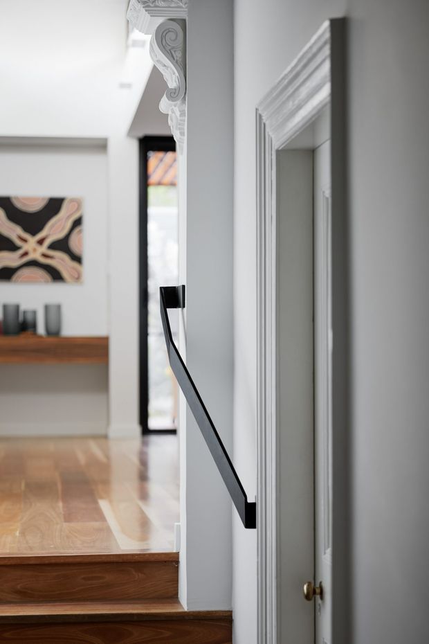 A refined interior hallway and stairwell with polished timber stairs and flooring, white walls, ornate decorative cornices and door trim, a slim black stair balustrade, and a built-in timber console shelf displaying dark vessels. A framed abstract artwork and a glazed exterior door are visible in the background.