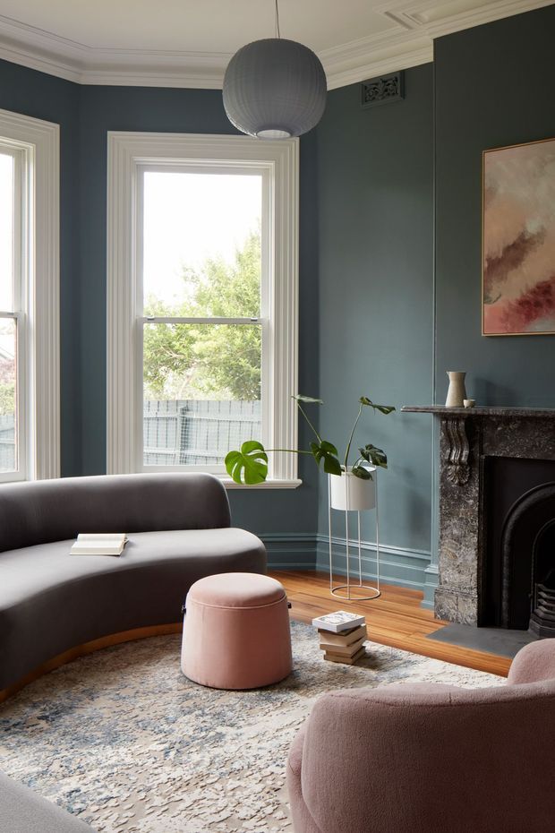 A refined living room with deep blue-green paneled walls, ornate white ceiling cornices, tall sash windows, and a dark stone fireplace with a black fire surround. Curved charcoal and blush upholstered seating surrounds a pink ottoman and patterned area rug, while a large spherical pendant light and potted monstera add sculptural accents. The room combines period architectural detailing with contemporary furnishings.