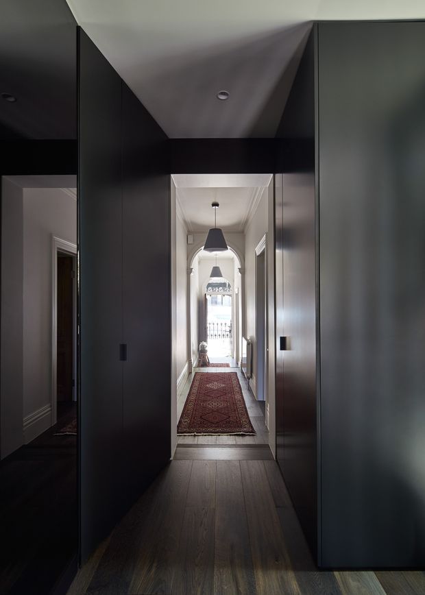 A narrow, moody residential hallway framed by tall, dark built-in cabinetry or wall panelling. The foreground has dark timber flooring, while a brighter traditional corridor beyond features an arched opening, ornate ceiling detailing, pendant lights, white trim, and a long patterned runner rug leading toward a glazed exterior door or window.