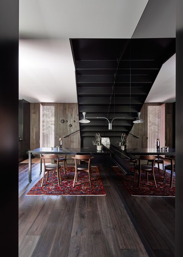 A dramatic, symmetrical dining area with a long dark dining table and wooden chairs arranged over richly patterned red rugs. Dark timber flooring and wood-panelled walls frame a large black sculptural overhead structure with integrated pendant lighting. Vertical timber screens, dark built-in joinery, mirrors, and carefully placed decorative branches create a refined, high-contrast contemporary interior.