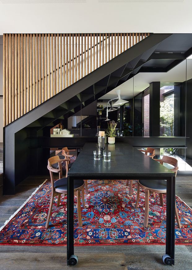 A contemporary open-plan dining and kitchen area beneath a dramatic black staircase. A large dark dining table with timber-and-upholstered chairs sits on a richly patterned red, blue and cream rug. Black built-in cabinetry and shelving occupy the area beneath the stairs, while vertical timber screening forms a striking upper-level balustrade. Full-height glazing brings in abundant natural light and views of greenery.