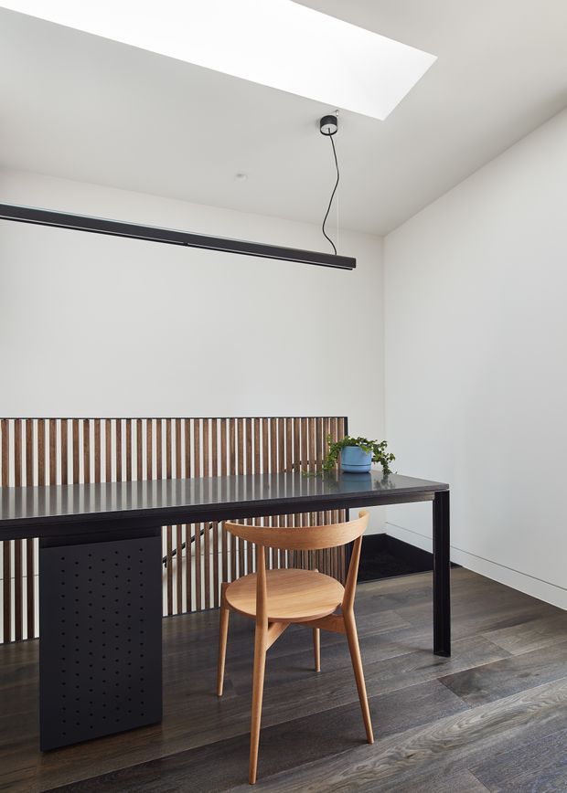 A minimalist dining or work area with a long black table, a sculptural timber chair, dark timber-look flooring, and a vertical timber slat screen along the rear edge. White walls and ceiling create a bright, restrained interior. A large rectangular ceiling opening or skylight and a suspended linear black light add architectural interest, while a small potted plant sits on the table.