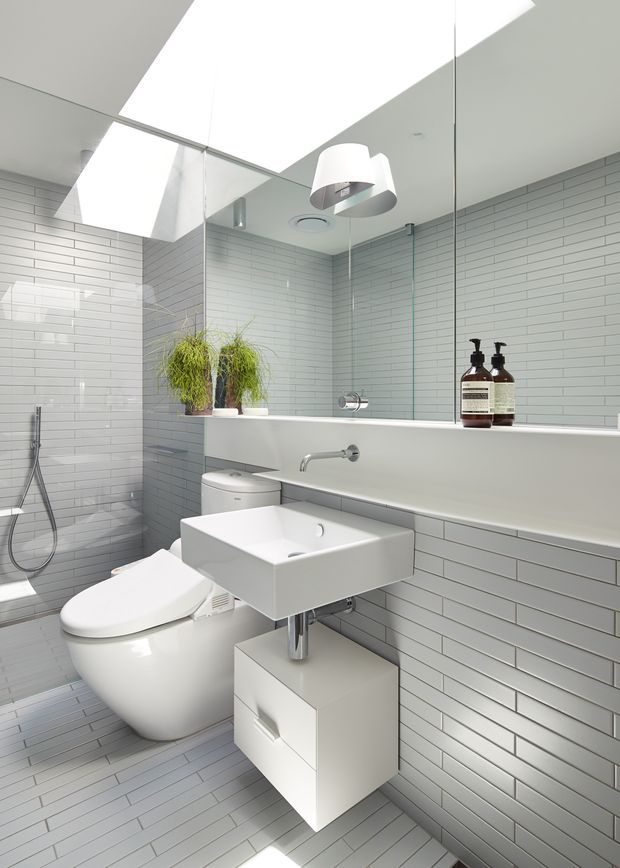 Bright, compact contemporary bathroom with full-height mirrored walls, pale grey-white rectangular tiled walls and floor, a wall-mounted square basin with white vanity cabinet, wall-hung toilet, and a narrow built-in bathtub or ledge beneath the mirror. A glass shower enclosure, chrome fixtures, potted greenery, and pendant lighting complete the clean minimalist fit-out.
