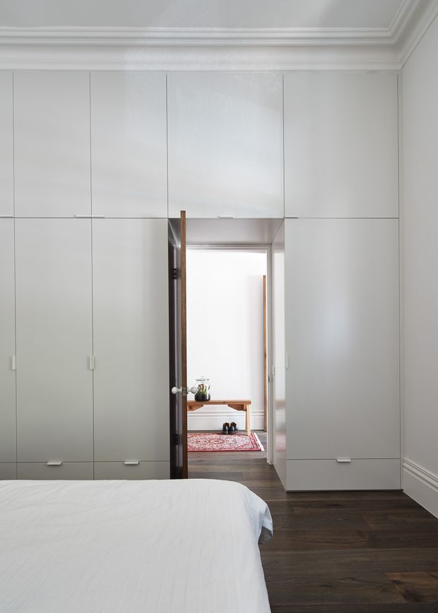 A bright, refined bedroom with a partially visible bed in the foreground, dark timber flooring, and extensive floor-to-ceiling white built-in cabinetry covering the walls around a central doorway. The room has ornate white crown moulding and skirting boards. Through the doorway, a small hallway or adjacent space with a console table and patterned rug is visible.
