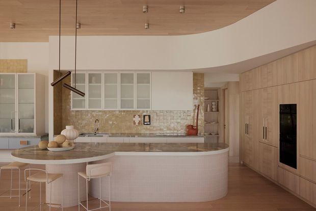 A spacious, softly curved contemporary kitchen with a large rounded island and integrated breakfast bar, pale ribbed cabinetry, full-height timber joinery, and a warm timber ceiling. The island has a stone benchtop and light pink-toned vertical tile cladding, while the rear wall features a textured beige mosaic tile splashback and pale glazed display cabinetry. Minimal black pendant lighting and integrated appliances complete the refined, warm-toned interior.