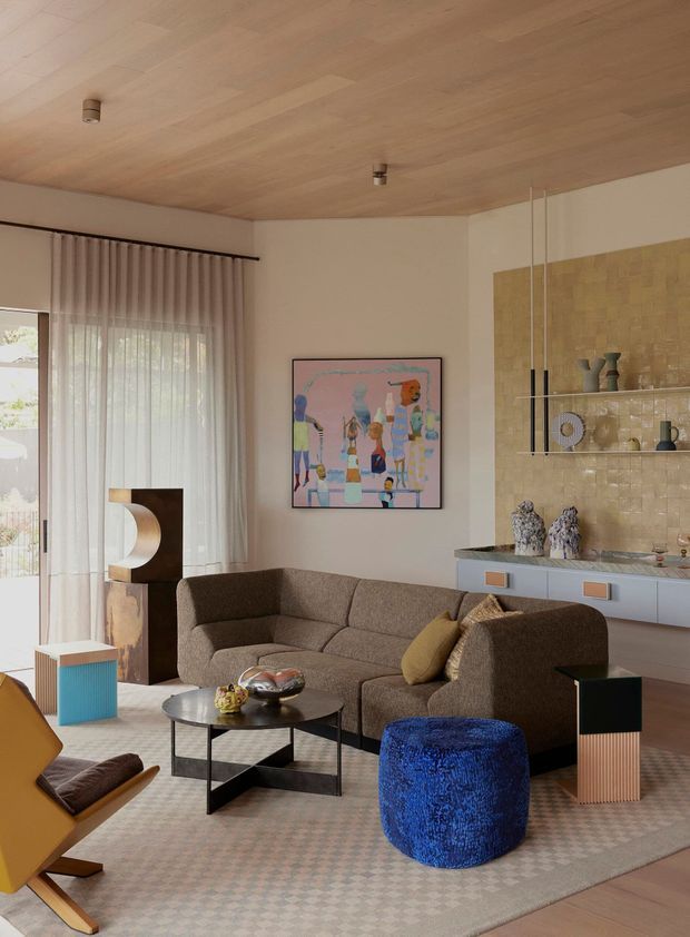 A refined living room with a warm timber-lined ceiling, pale neutral walls, full-height sheer curtains, and a large brown sectional sofa arranged around a low black coffee table. The room features a built-in pale blue-grey display cabinet and wet-bar style joinery with a textured gold-toned tile backsplash and open shelving. Sculptural lamps, colourful artwork, patterned rug, and vivid blue ottoman add contemporary artistic accents.