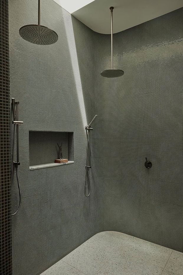 A contemporary walk-in shower room clad floor-to-ceiling in tiny dark grey mosaic tiles. The curved corner walls contain two large round rainfall showerheads, a handheld shower, recessed shelf, and dark metal fixtures. The floor is a light speckled stone or terrazzo-like finish, creating a restrained, moody, monolithic bathroom interior.