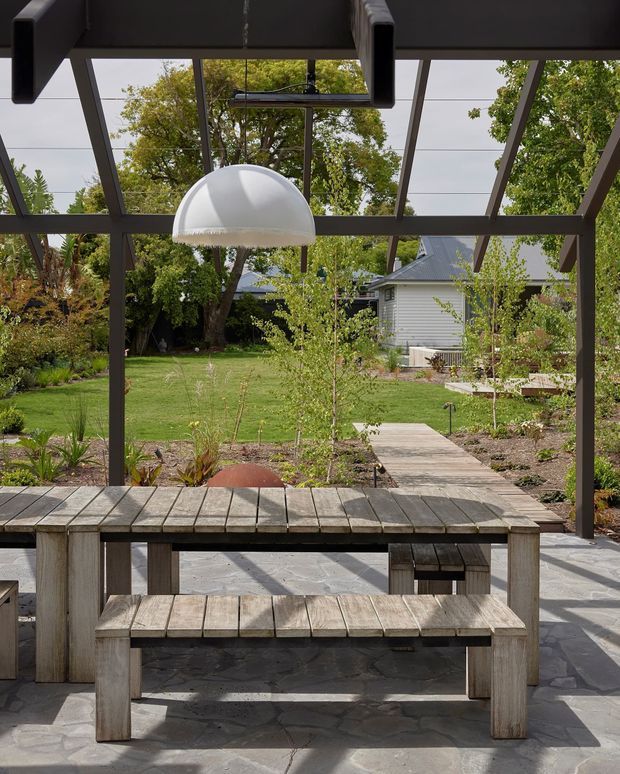 A covered outdoor dining and entertaining area with a rustic timber picnic-style table and benches beneath a dark timber pergola or glazed garden pavilion. A white pendant light hangs overhead, while a paved terrace leads through landscaped garden beds toward a lawn and a weatherboard outbuilding. The scene has a relaxed, natural residential garden setting with strong indoor-outdoor connection.