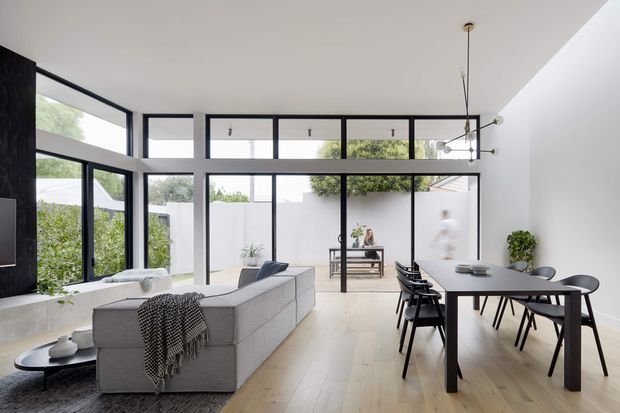 Bright, open-plan contemporary living and dining area with double-height glazing, black-framed windows, pale timber flooring, and a restrained neutral palette. A large modular sofa faces a dark feature wall, while a long dining table with black chairs occupies the foreground. The rear glazing opens visually to a small planted courtyard, and a sculptural linear pendant hangs above the dining area.