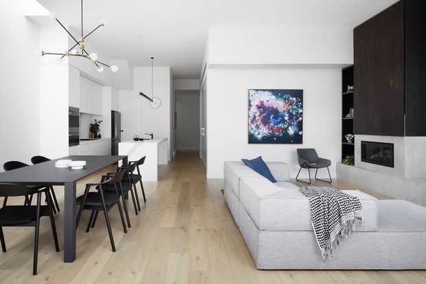 Bright open-plan contemporary interior combining a dining area, kitchen and living room. The space features pale timber flooring, white walls and cabinetry, a large light-grey sectional sofa, a dark dining table with black chairs, sculptural pendant lighting, and a dark feature wall containing a minimalist fireplace. Abstract artwork and restrained furnishings create a clean, refined atmosphere.