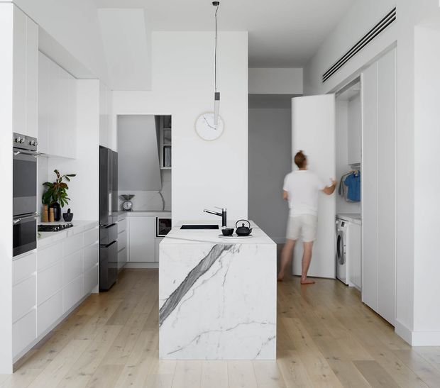 Bright, minimalist open-plan kitchen with white full-height cabinetry, integrated appliances, pale timber flooring, and a compact marble-look island with an undermount sink and black tapware. A small pantry or utility area is visible at the rear, while an open laundry or wardrobe space with a washing machine and hanging clothes is visible to the right. A simple pendant light and clean white finishes create an airy contemporary interior.