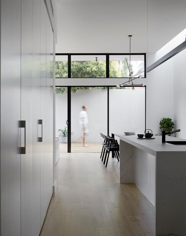 A bright, minimalist kitchen and dining area viewed along a long circulation space. The room features full-height white cabinetry with dark vertical handles, a large pale stone island with an integrated black tap and sink, black-framed glazing opening to a private courtyard, pale timber flooring, and a sculptural linear pendant light. A compact dining setting sits beside the island, with abundant natural light and greenery visible outside.