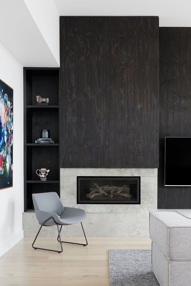 Contemporary living room featuring a dark vertically slatted timber feature wall with integrated shelving and a wall-mounted television. A minimalist linear fireplace is set into a pale stone or concrete surround, with light timber flooring, a grey lounge chair, pale upholstered seating, and a textured grey rug. The room has a restrained monochromatic palette with a colourful artwork at the edge of the frame.