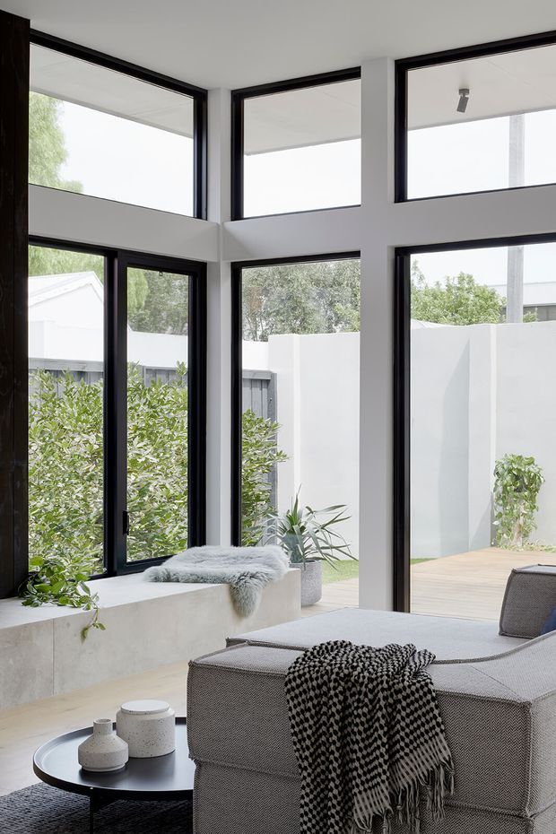 A bright, contemporary interior lounge with a large double-height glazed corner opening onto a private courtyard. Black-framed windows and doors contrast with white walls and concrete-style built-in bench seating. A neutral upholstered lounge chair, round black coffee table, and monochrome throw create a refined, minimalist atmosphere, with potted greenery visible inside and outside.