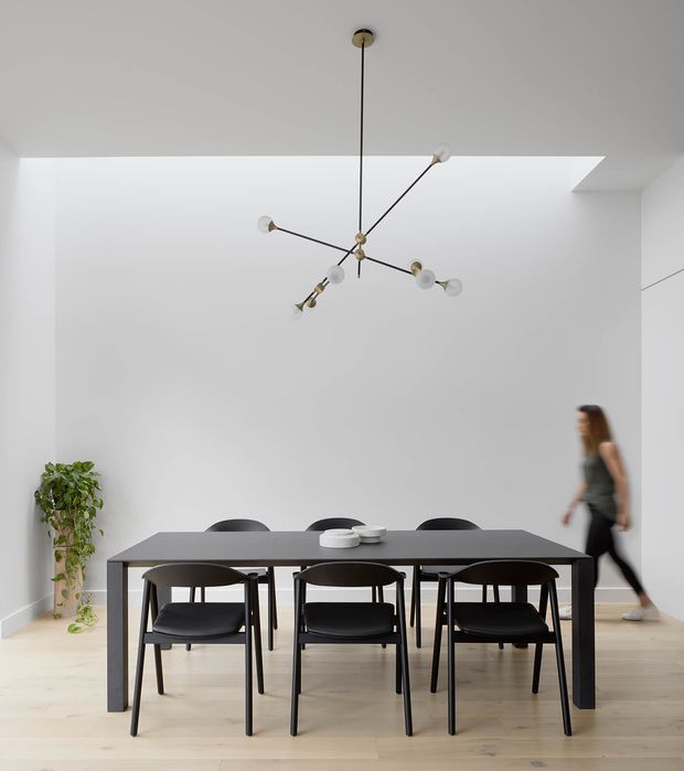 A minimalist dining room with a long dark rectangular dining table and six dark dining chairs arranged beneath a sculptural branching pendant light. The room has white walls, pale timber flooring, a recessed ceiling detail, and a small potted plant in the corner. A blurred person is walking along the right side.