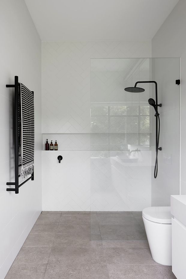 A bright, compact contemporary bathroom with a walk-in glass shower, black shower hardware, wall-mounted black towel warmer, white geometric-pattern wall tiles, and large grey floor tiles. A recessed wall niche holds small bottles, while a white toilet and vanity are visible on the right. The room has a clean, minimalist monochrome palette with subtle natural light through the shower glazing.