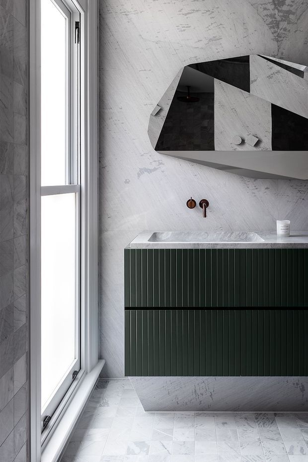 A contemporary bathroom vanity with a dark green vertically fluted wall-mounted cabinet, pale stone benchtop and integrated basin, brushed bronze wall-mounted tapware, and a large angular sculptural mirror. The bathroom features pale grey marble-look wall and floor finishes, a tall white-framed window, and a monochromatic palette accented by deep green and bronze.