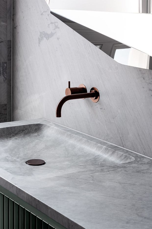 Close-up of a refined bathroom vanity featuring a wall-mounted dark bronze tap, integrated grey stone basin and matching stone splashback. A narrow mirror or reflective strip runs above the basin, while dark green vertical cabinetry is visible below. The composition is minimalist and material-focused, with soft natural light and subtle tonal contrast.
