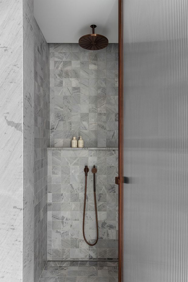 Compact contemporary shower enclosure with grey veined marble-look wall and floor tiles, a recessed tiled niche with toiletries, a large round bronze overhead shower, matching handheld shower fittings, and a ribbed translucent glass door in a timber-toned frame.