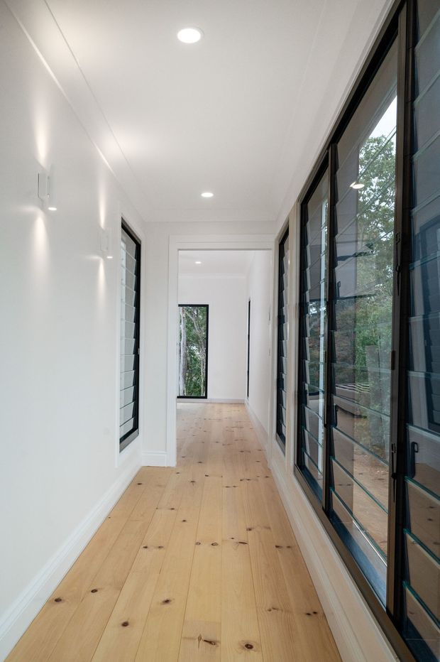A bright, narrow interior hallway with pale natural timber plank flooring, white walls and ceiling, recessed downlights, and extensive black-framed glazed louvre windows along the right side. Additional black-framed windows and a view through to greenery at the far end provide natural light and visual connection to the outdoors.