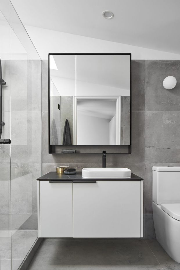 A compact contemporary bathroom with large grey concrete-look wall and floor tiles, a frameless glass shower enclosure, floating white vanity with a dark benchtop, white vessel basin, black tapware, and a large black-framed mirrored cabinet. A toilet is visible on the right, with minimalist wall lighting and clean monochromatic finishes.