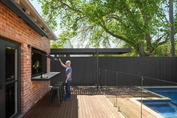 A sunny outdoor entertaining area beside a red brick house, featuring a timber deck, built-in outdoor bar with stools, dark privacy fencing, a retractable roof or awning, and a glass pool fence enclosing a swimming pool. Mature trees surround the space.