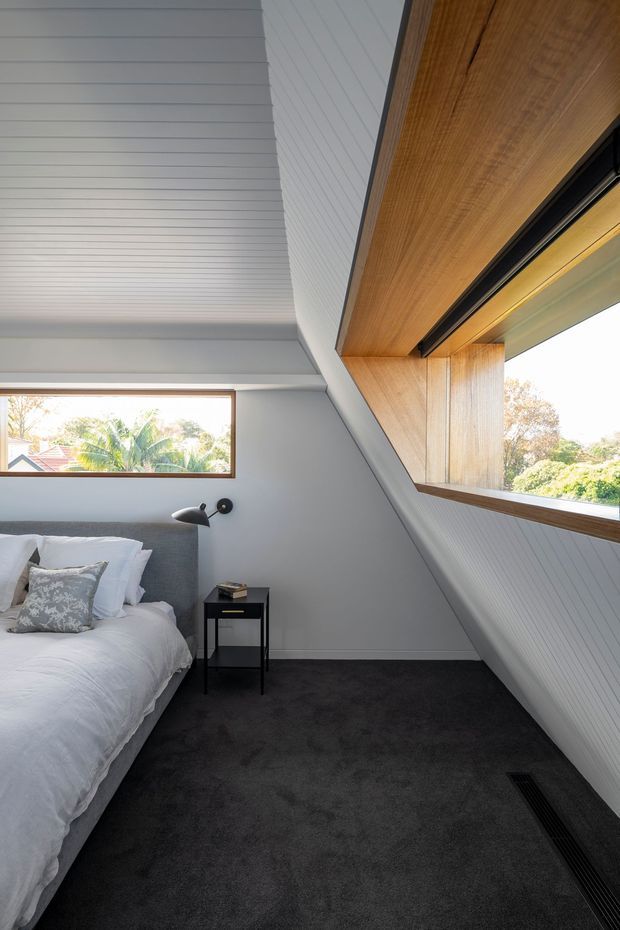 A compact contemporary bedroom beneath a sloped ceiling, featuring white linear wall and ceiling panelling, dark charcoal carpet, a partially visible upholstered bed with white bedding, a small black bedside table, and minimalist black wall lighting. Large horizontal windows with warm timber reveals bring in natural light and views of trees and neighbouring rooftops.