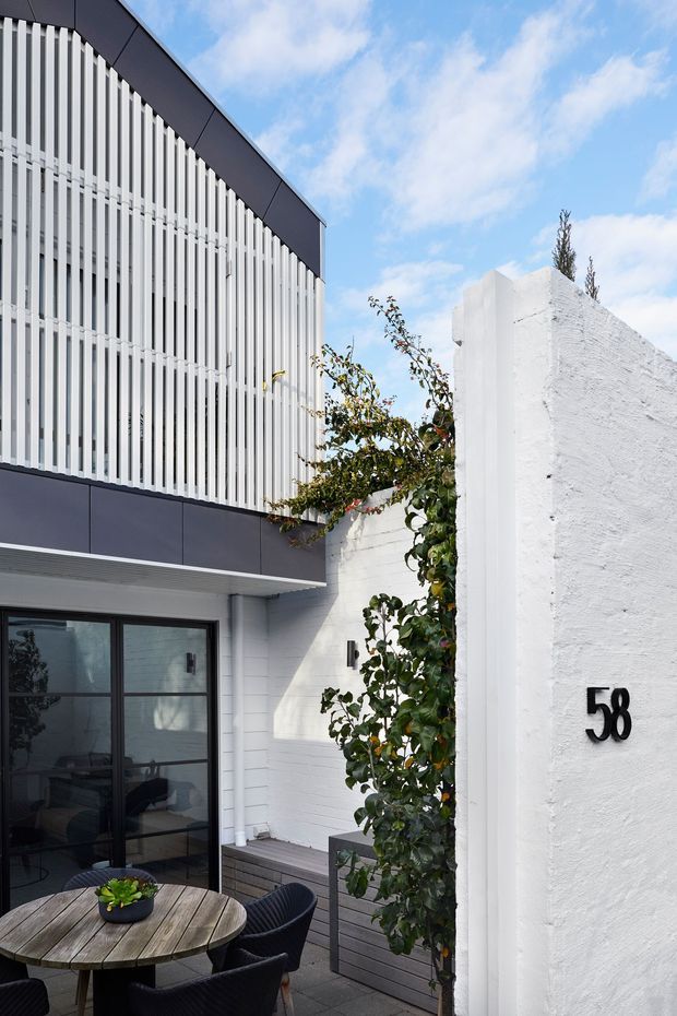 A bright contemporary courtyard entrance viewed from outdoors. The two-storey facade combines dark charcoal cladding panels with extensive white vertical screening, dark-framed glazed doors, and white painted masonry walls. A small paved outdoor dining area with a round timber table and chairs sits beside a planted trellis, while a white rendered boundary wall displays the house number 58.