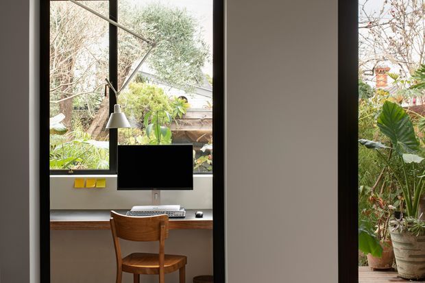 Compact home office nook with a built-in timber desk beneath large black-framed windows. A computer monitor, desk lamp, keyboard and wooden chair create a functional workspace, while lush planting is visible outside. The room has a restrained, contemporary feel with neutral walls and dark window framing.