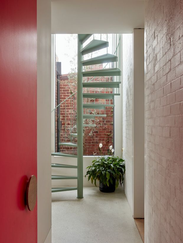A narrow, light-filled interior passage with a sculptural pale green spiral staircase beside full-height glazing. The space features a polished light concrete floor, exposed painted brick walls, a vivid red door with a timber handle, and a potted plant. A small brick courtyard is visible through the glazing.