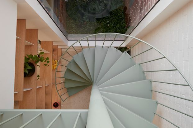 A downward-looking view of a sculptural spiral staircase with pale grey metal treads and a curved wire balustrade. The staircase sits within a narrow, light-filled interior void, bordered by white-painted brick walls and warm timber built-in shelving with indoor plants. A glazed overhead opening reflects or reveals greenery, adding contrast to the restrained material palette.