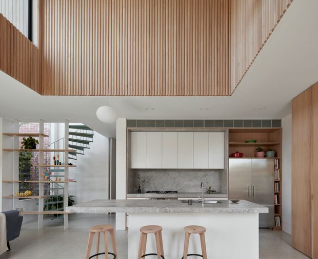 Bright contemporary kitchen with a large pale stone island, integrated white cabinetry, timber veneer storage, open shelving, and three timber stools. A textured grey stone splashback frames the cooktop, while vertical timber battens form a dramatic ceiling feature above the open-plan space. Glazed shelving and a sculptural staircase are visible to the left.