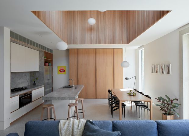 Bright open-plan kitchen, dining, and living area with a contemporary minimalist fit-out. The kitchen features white flat-panel cabinetry, a grey stone island and benchtop, and integrated appliances. Warm timber cabinetry and a timber slatted recessed ceiling create a strong feature element, while a compact dining table, black chairs, pendant lights, and a blue sofa occupy the adjoining spaces.