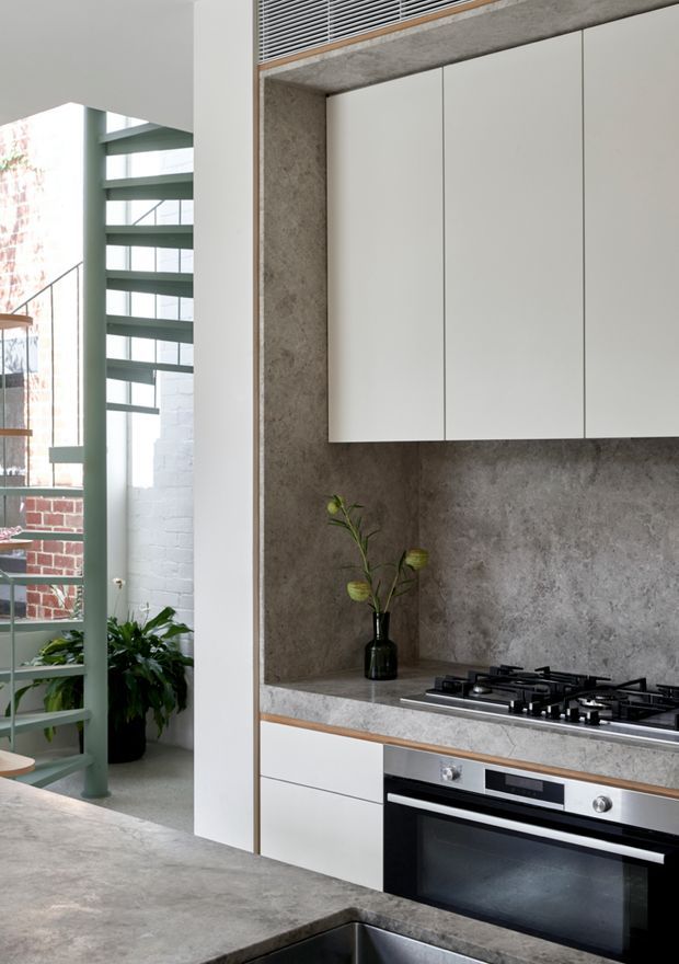 Contemporary kitchen with minimalist white flat-panel cabinetry, a grey stone or concrete-look splashback and benchtop, integrated stainless steel appliances, and a black gas cooktop. A concrete-look island with inset sink occupies the foreground. The kitchen opens toward a bright industrial-style stairwell with pale green metal railings, exposed brick, and large glazing.