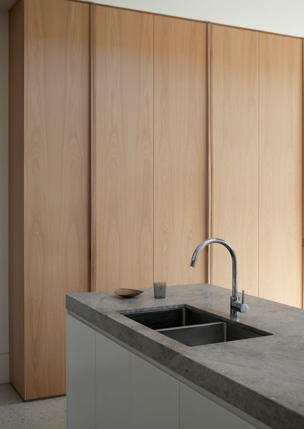 A close-up view of a minimalist kitchen island with a large grey stone countertop, integrated dark double-bowl sink, and chrome gooseneck tap. The island has pale grey flat-panel cabinetry, while full-height warm timber veneer cabinetry forms the backdrop. The composition is clean and understated with soft natural lighting.