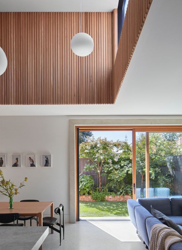 Bright open-plan interior combining a dining area and living room, viewed toward a lush private garden through large timber-framed glazing. A dramatic ceiling feature clad in closely spaced vertical timber battens contains spherical pendant lights, while the lower portion shows a timber dining table, black dining chairs, a sectional sofa, framed wall art and a pale stone or concrete kitchen bench in the foreground. The room has a clean contemporary character with strong indoor-outdoor connection and abundant natural light.