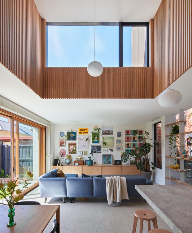 Bright contemporary living room with a large blue sectional sofa, timber-framed glazing, a gallery wall of artwork and photographs, indoor plants, open shelving, and a substantial stone-topped kitchen island or counter. A dramatic double-height ceiling is lined with vertical timber battens and includes a large clerestory window and suspended spherical pendant lights.