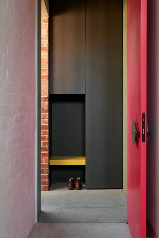 A narrow architectural entryway featuring a tall dark built-in storage unit with open shelving, black interior surfaces, and a mustard-yellow shelf. Exposed red brick is visible beside the joinery, with pale painted brick walls, a vivid pink-red door, and a grey concrete floor. The composition highlights bold colour blocking, integrated storage, and contrasting raw and painted masonry.