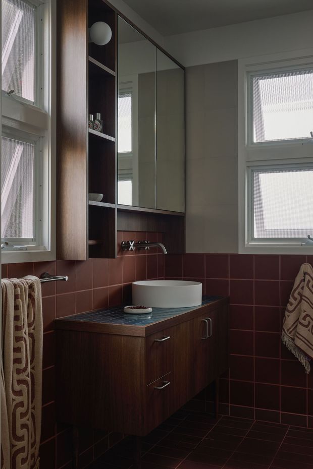 A compact bathroom with a dark timber floating vanity, white vessel basin, wall-mounted chrome tapware, mirrored upper cabinetry, and open shelving. Deep burgundy-red square tiles cover the walls and floor, while white-framed textured-glass windows provide filtered natural light. Patterned towels add a small decorative accent.