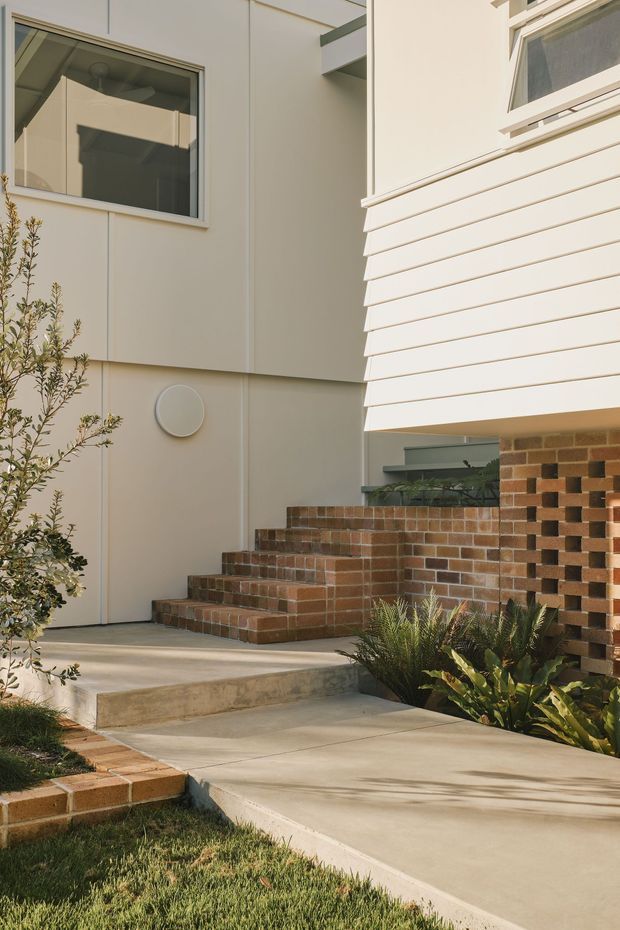 A close exterior view of a contemporary residential entry courtyard. Pale rendered or panelled walls with simple rectangular windows sit above a concrete walkway and stepped brick paving. A projecting upper volume is clad in horizontal white weatherboards, while a perforated brick privacy wall and brick steps define the entry. Low ornamental planting borders the path.