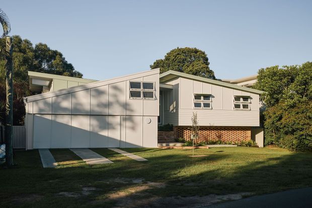 Exterior view of a raised, low-profile residential house with pale cladding, broad roof overhangs, a projecting garage or carport volume, horizontal windows, and a partially screened lower level. The home sits in a leafy suburban garden with a simple lawn and concrete driveway strips.