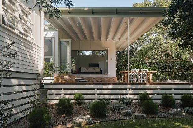 Exterior view of a contemporary single-storey home addition or covered outdoor living area. The image shows a pale horizontal weatherboard facade, a broad flat skillion-style roof with exposed painted rafters, open glazing and doors leading into a living room, and a timber-clad raised deck behind a horizontal slatted privacy fence. A small outdoor dining setting and landscaped planting sit beside the covered terrace, with mature trees beyond.